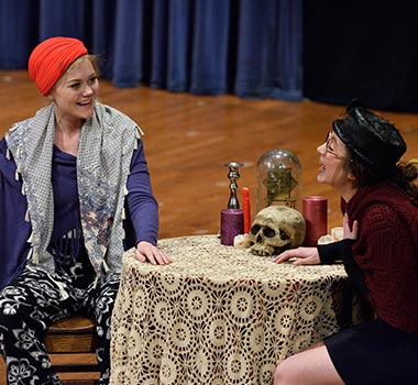 Young Playwrights Festival, Two people act on stage while sitting at a table with ornaments and skull. 